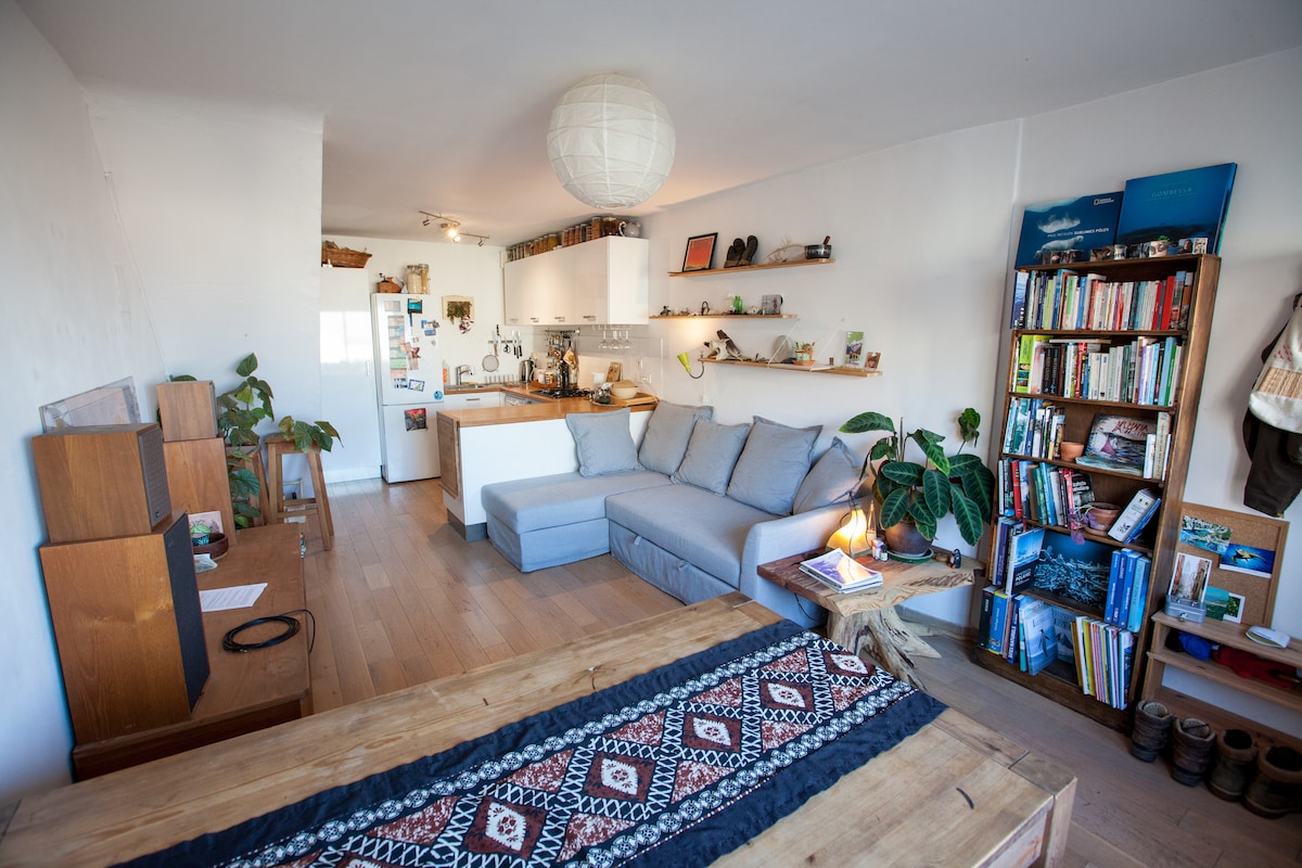 A bright living space features a comfortable gray sofa and a rustic wooden table adorned with a decorative runner. Shelves display books and decor, while plants add a touch of nature. A modern kitchen is visible in the background, enhancing the open feel.