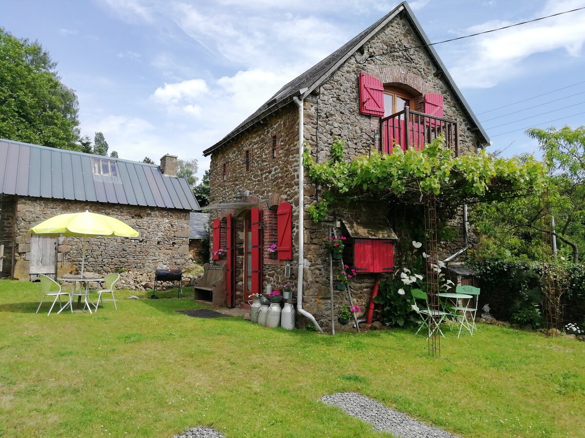The stone barn features vibrant red shutters and a small balcony overlooking the grassy courtyard. A yellow umbrella provides shade for outdoor seating which includes tables and chairs. Lush greenery and flowering plants enhance the inviting exterior. A grilling area is also visible.