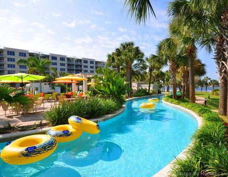 A winding lazy river pool is surrounded by palm trees and lush greenery. Bright yellow inner tubes float in the sparkling water, which is bordered by lounge chairs and umbrellas for shade. The vibrant flowers and plants create a tropical ambiance.