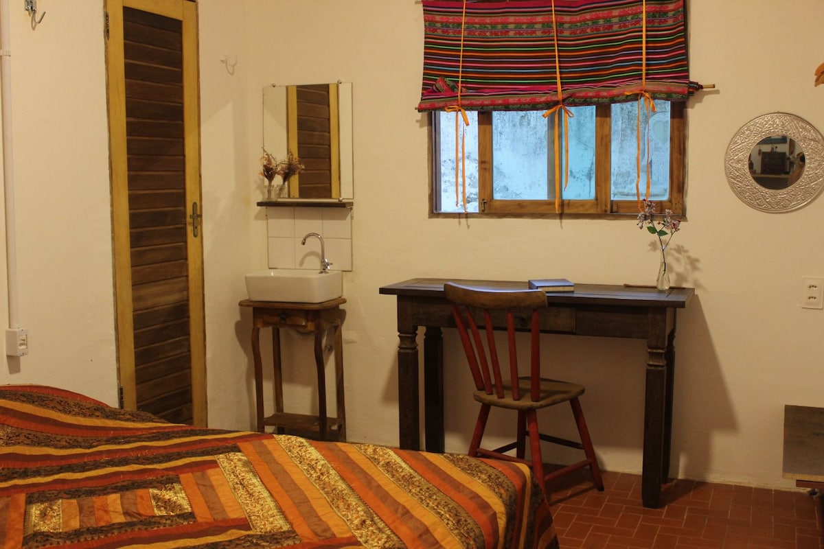 An inviting bedroom features a spacious bed adorned with a multicolored striped blanket. A wooden desk and red chair provide a workspace, while a small mirror and sink add functionality. A window with decorative curtains allows for natural light to fill the room.