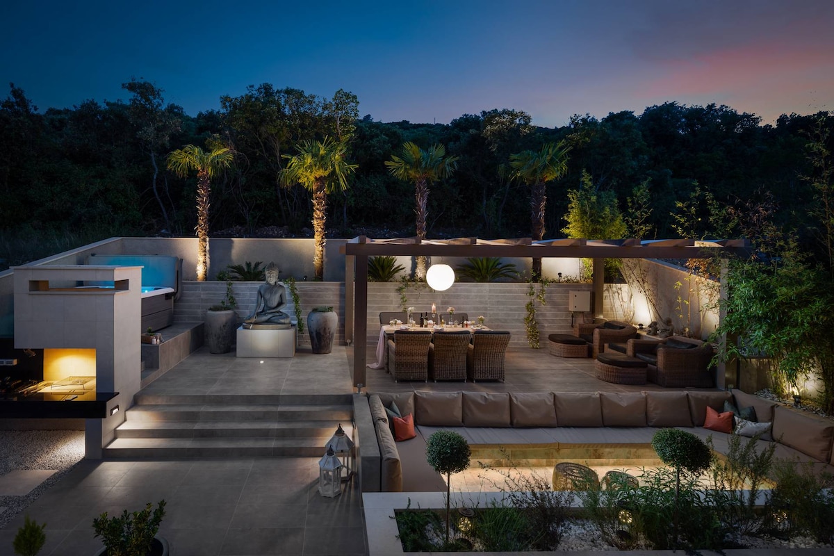 An outdoor area is showcased at dusk, featuring a dining table and multiple lounge settings beneath a wooden pergola. A large Buddha statue is positioned centrally, surrounded by lush greenery and illuminated elements. The Jacuzzi and grill are visible, contributing to a serene atmosphere.