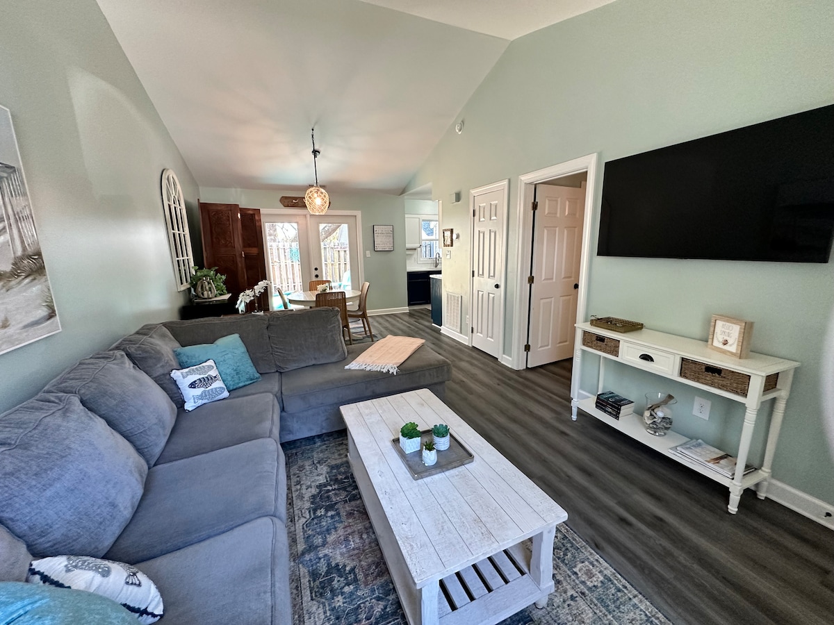 A spacious living area is showcased, featuring a large sectional sofa in soft grey and a rustic coffee table. Natural light floods the room through a nearby window, highlighting the light green walls and modern decor. A doorway leads to an adjacent dining space.