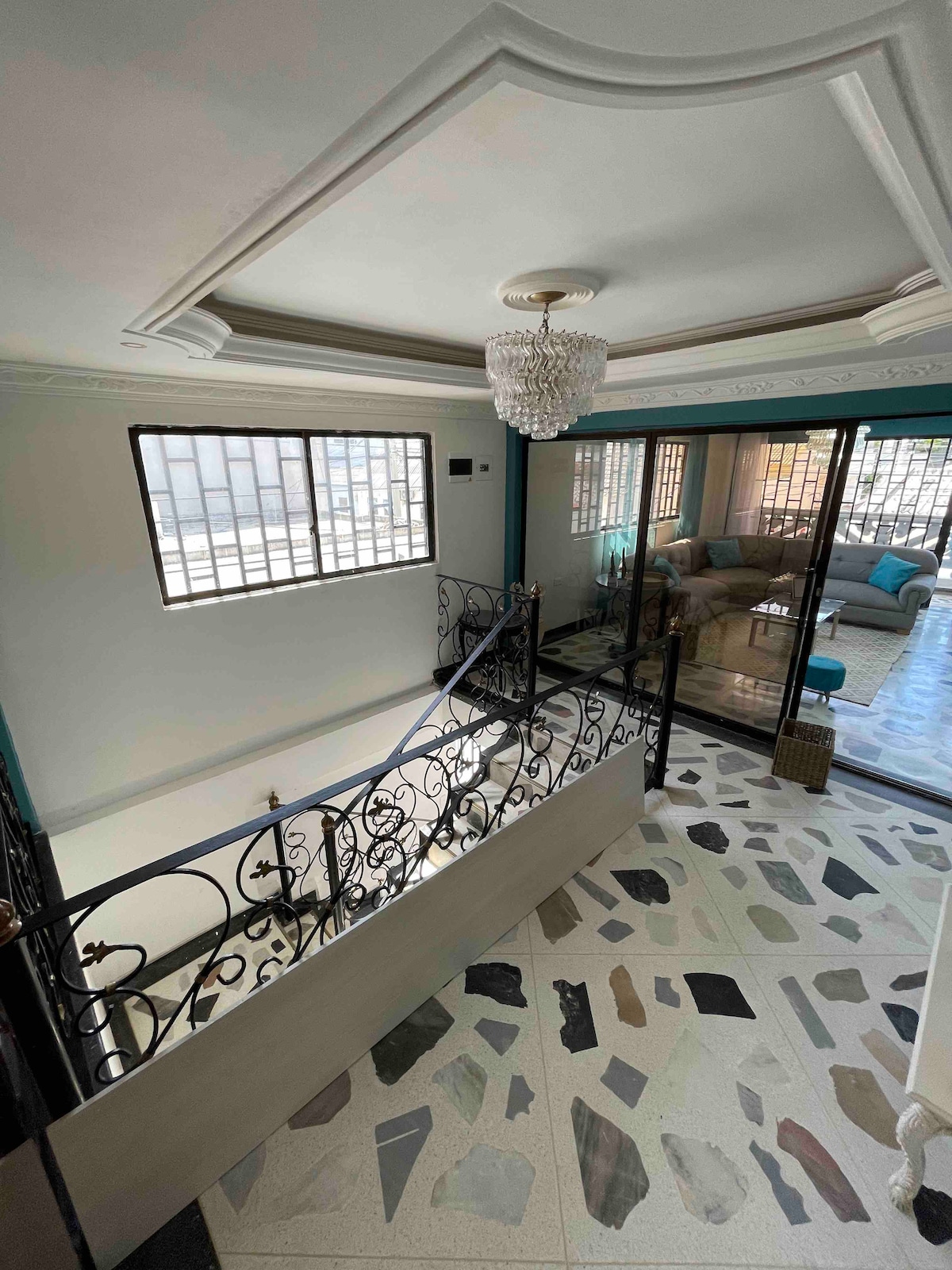 A spacious upper landing features elegant wrought iron railings, leading to a well-lit area with large windows offering views of the surroundings. The floor is adorned with an array of colorful tiles, while a chandelier adds a touch of sophistication.