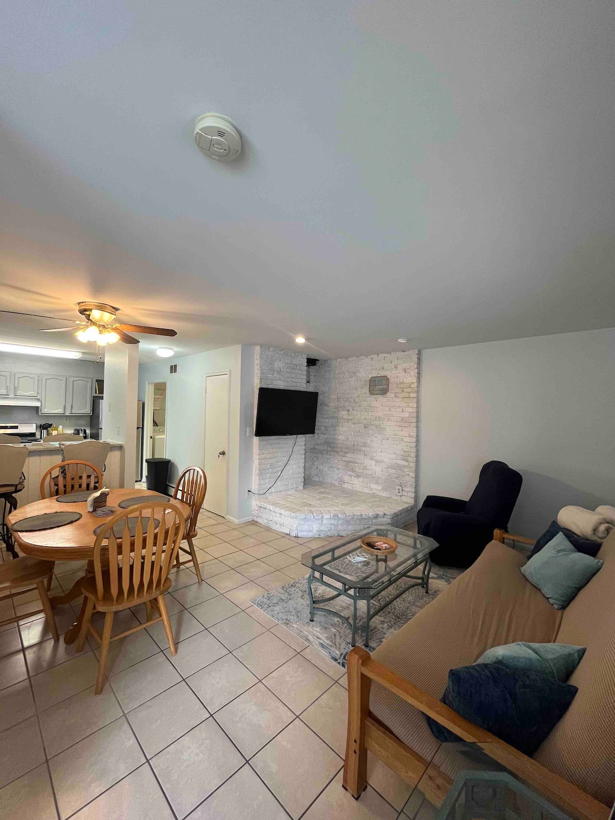 The living area features a light-colored tile floor with a cozy futon and a recliner. A round dining table with wooden chairs accommodates four. A TV is mounted on the wall, and natural light illuminates the space from nearby windows.