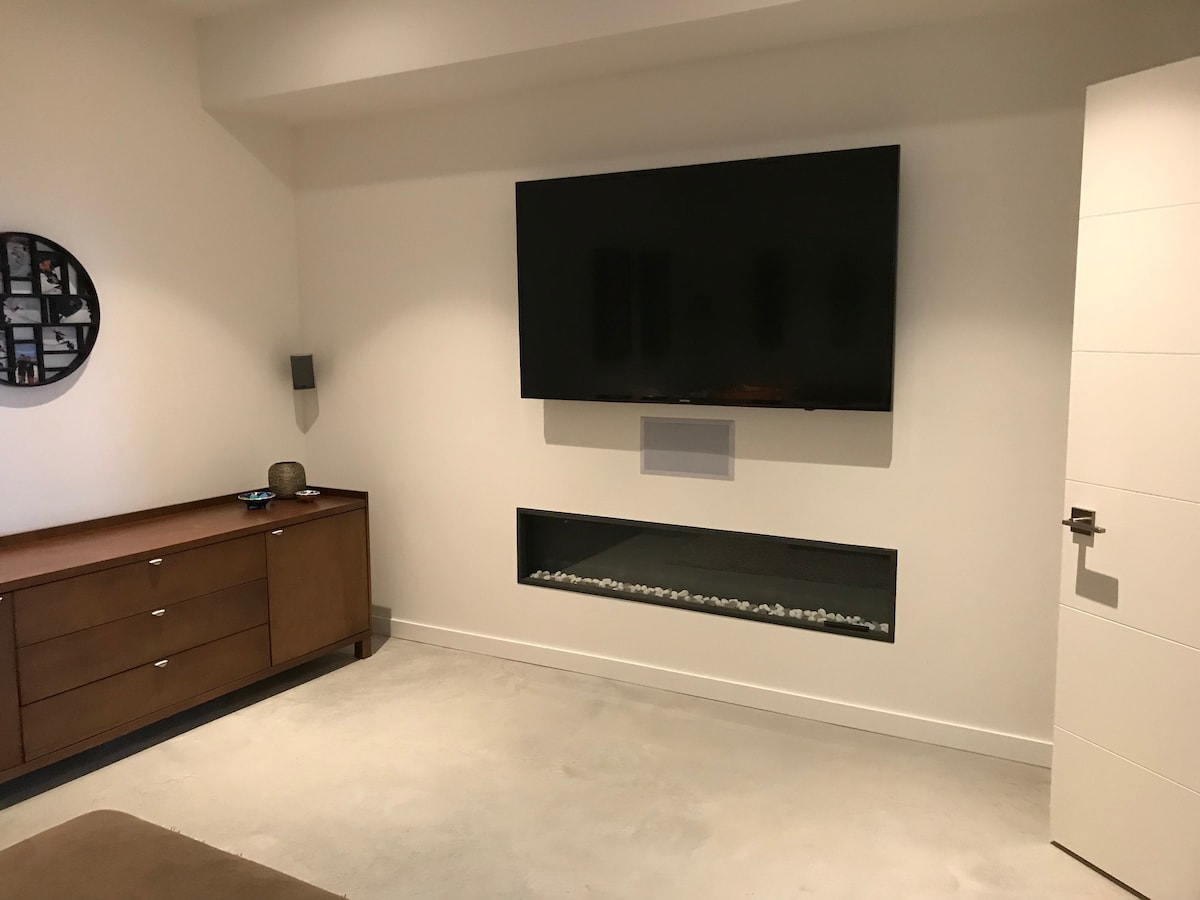 A modern lounge area features a large, wall-mounted television above a low entertainment unit made of rich wood. An electric fireplace with glass pebbles is built into the wall, adding a contemporary touch to the space.