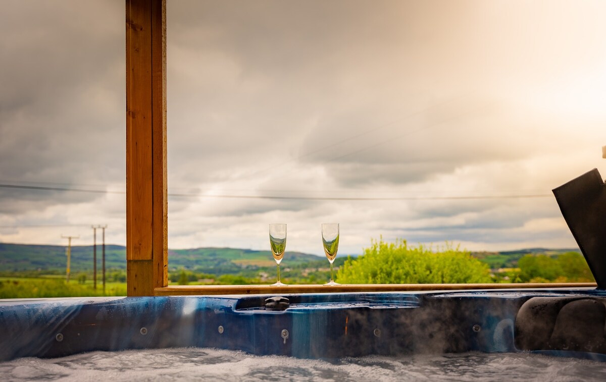 A private hot tub is seen with a scenic view of the surrounding landscape. Two champagne flutes rest on the edge of the tub, with steam rising gently in the cooler air. The sky is overcast, providing a serene backdrop for relaxation.