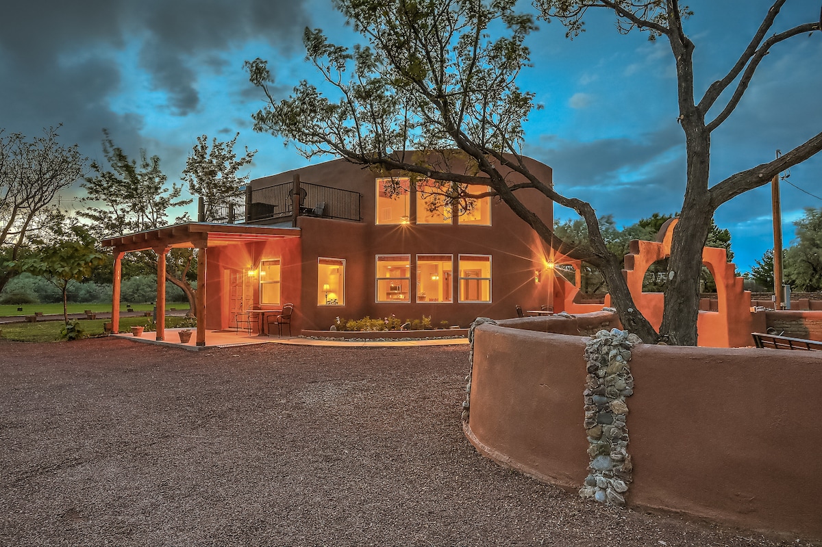 The exterior of the southwestern home is highlighted by soft evening light, showcasing its rounded architecture and vibrant earth-toned walls. Surrounding trees and a gravel pathway accentuate the serene environment, while large windows illuminate the inviting interior.