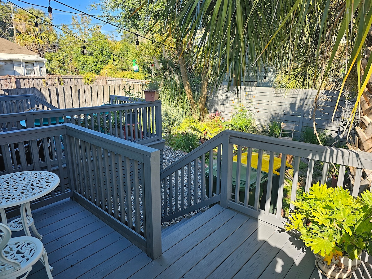 A wooden deck is accessed by a set of stairs, leading to a lush garden area. Casual seating, including a small round table and chairs, is visible. Surrounding greenery creates a vibrant, natural setting, accented by strung lights overhead, inviting relaxation in the outdoor space.
