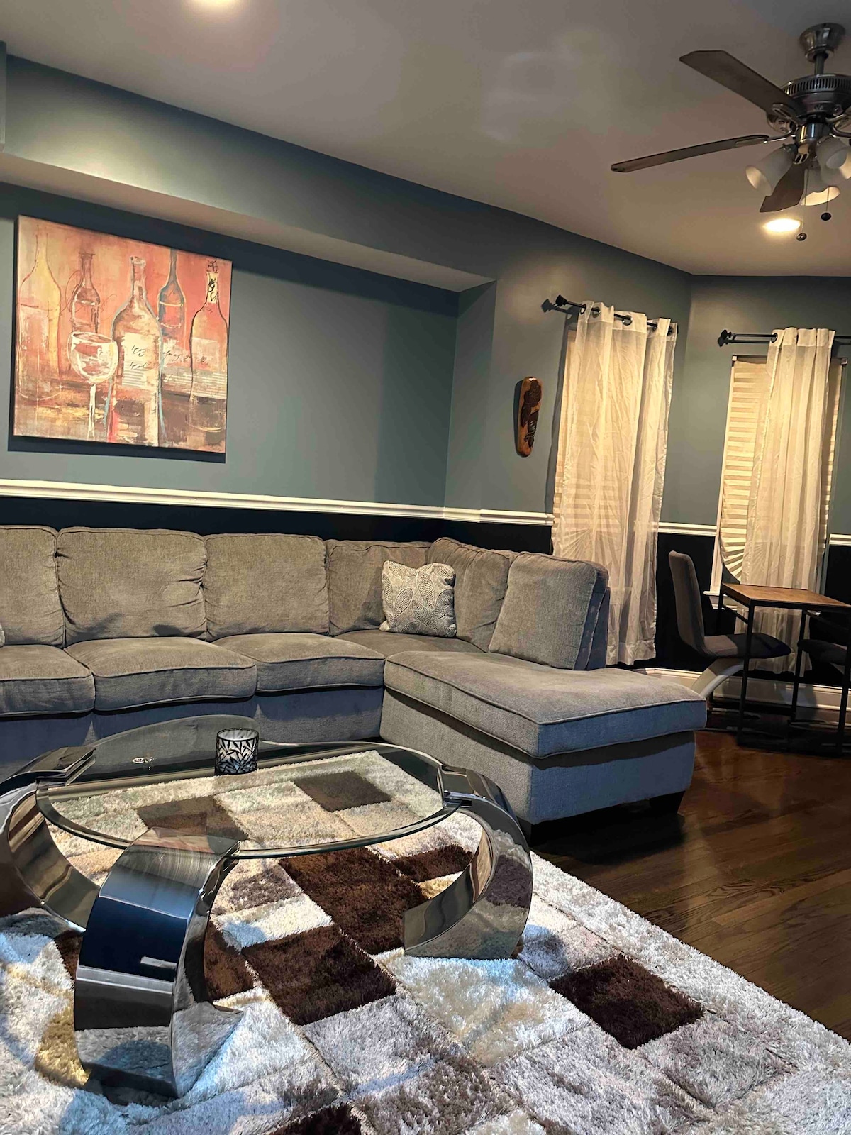 A spacious living area is furnished with a large sectional couch and a patterned area rug. A glass coffee table sits at the center, surrounded by neutral tones. Light filters in through sheer curtains, with a tasteful wall art piece depicting wine bottles enhancing the ambiance.