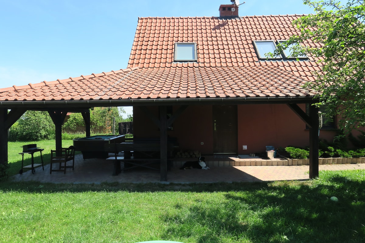 A two-story house is featured with a charming red tiled roof and a covered porch area. The outdoor space includes a grassy lawn, a barbecue grill, and seating arrangements. Trees enhance the natural surroundings, providing a peaceful setting for relaxation.