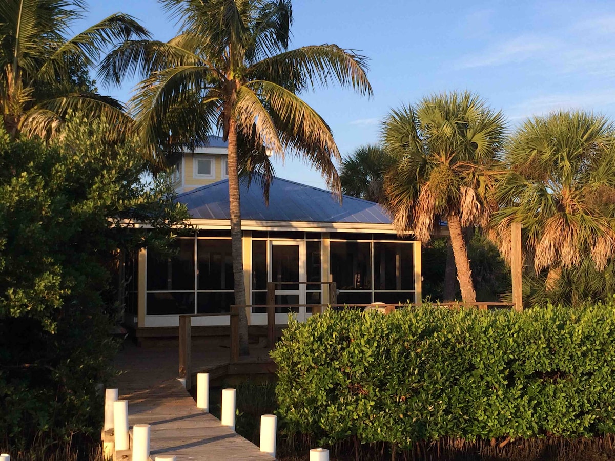 A welcoming guest house is surrounded by tropical greenery and palm trees. The structure features a screened porch that provides views of the landscaped area. A wooden walkway leads from the water to the house, enhancing the connection to nature.