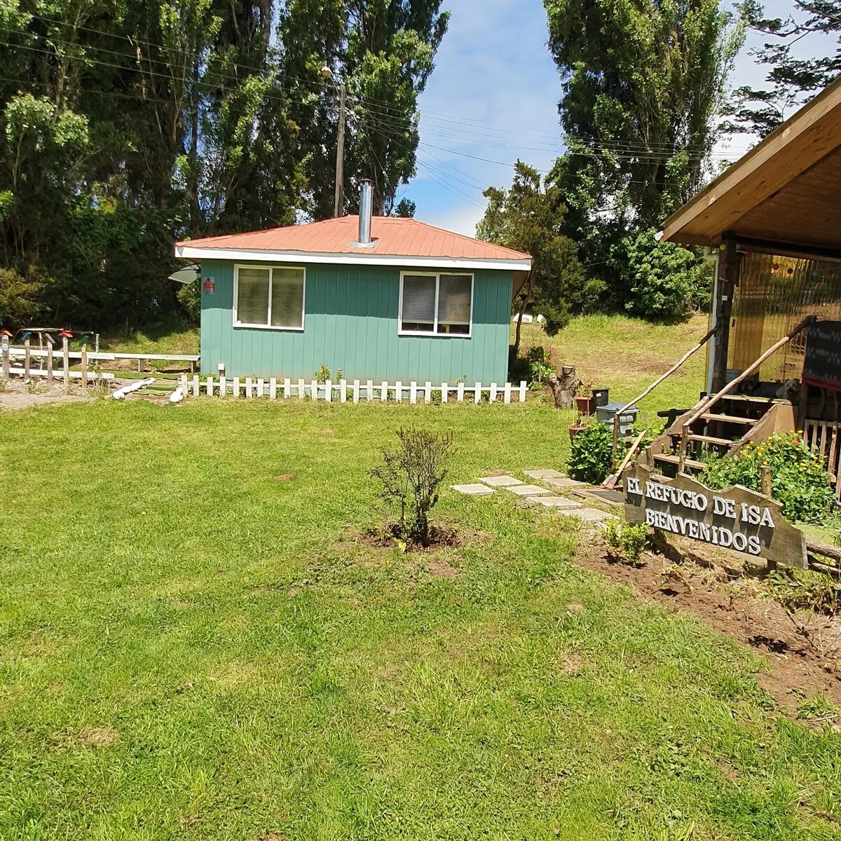 A charming green cabin is set among lush grass, featuring a red roof and white picket fence. The sign ‘El Refugio de Isa Bienvenidos’ is positioned near the entrance, with trees providing a natural backdrop to the tranquil surroundings.