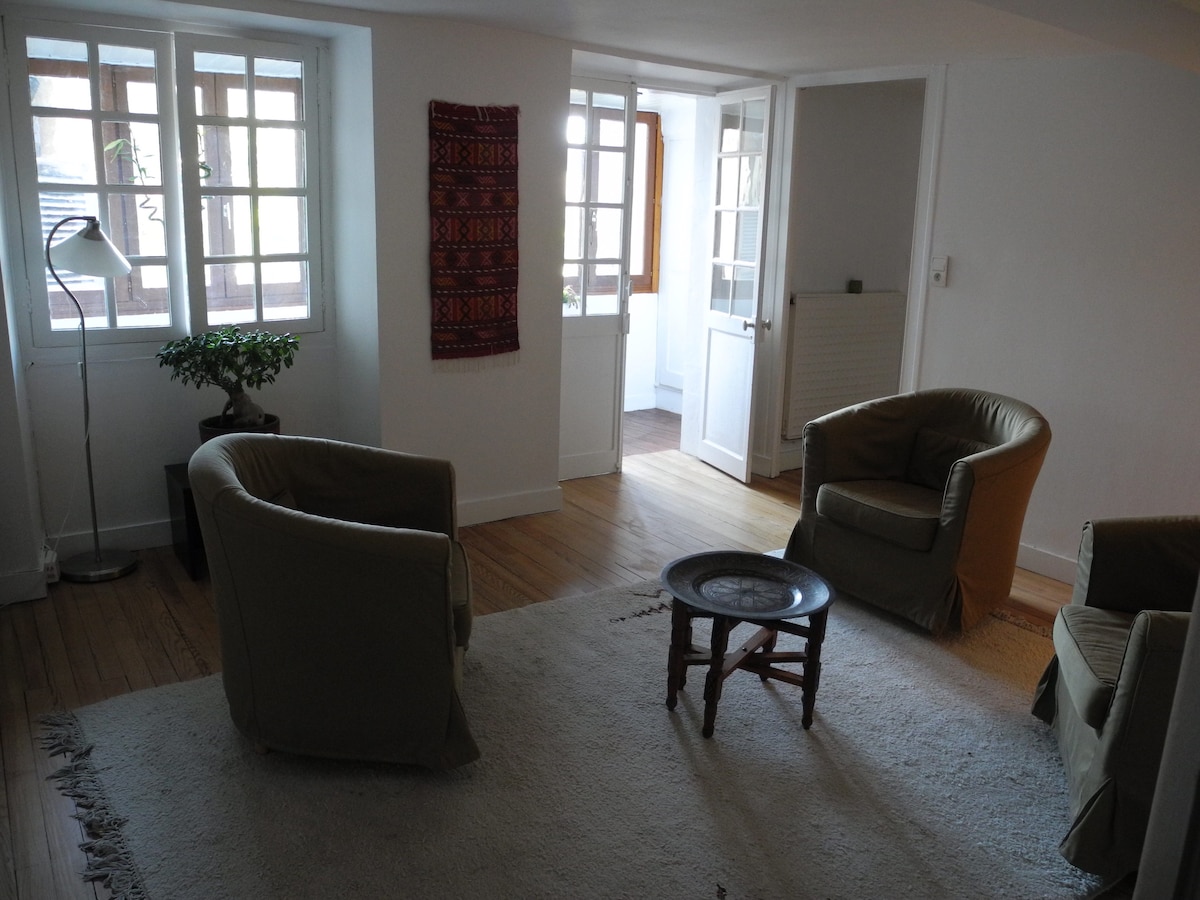 A cozy living area is highlighted by two comfortable armchairs and a small wooden table. Natural light filters in through large windows, illuminating the soft area rug. A decorative wall hanging adds a touch of character, while a potted plant contributes to the serene atmosphere.