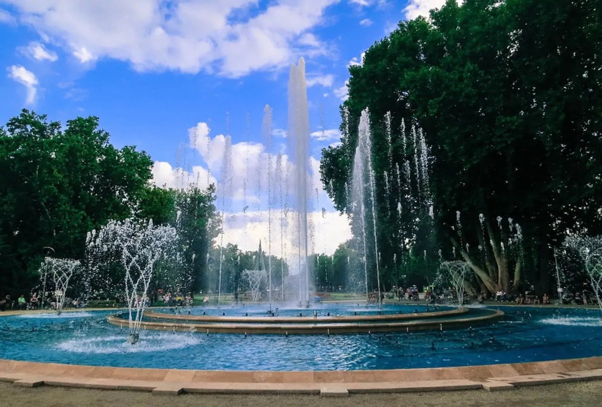 A large fountain is centrally featured, with water shooting upwards and creating an array of smaller splashes. Surrounding greenery enhances the serene environment, while a blue sky with scattered clouds provides a bright backdrop. Benches and people enjoying the park are visible in the distance.