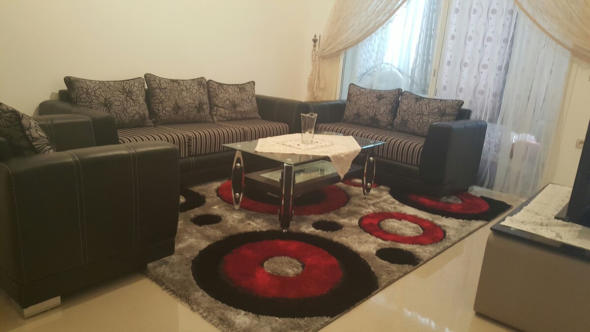 A comfortable seating area is arranged with two dark sofas and a glass coffee table in the center. A patterned area rug features bold red and black circles, while natural light filters through sheer curtains, softly illuminating the space. Decorative cushions add a touch of style.