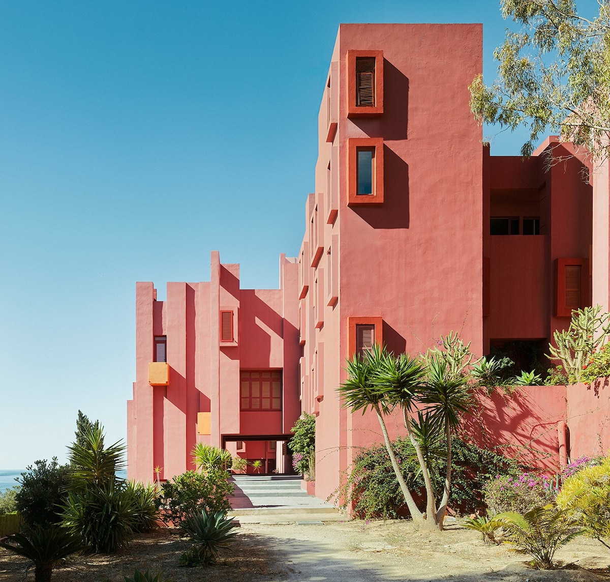 A striking candy-pink building with unique offset windows, designed by Ricardo Bofill, is contrasted against a bright, blue sky