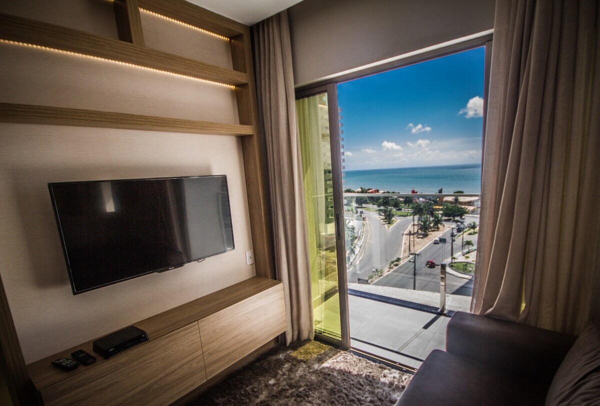 A modern living area features a large flat-screen TV mounted on a wall unit. A expansive window provides a view of the ocean and the surrounding landscape. Soft curtains frame the window, allowing natural light to fill the room.