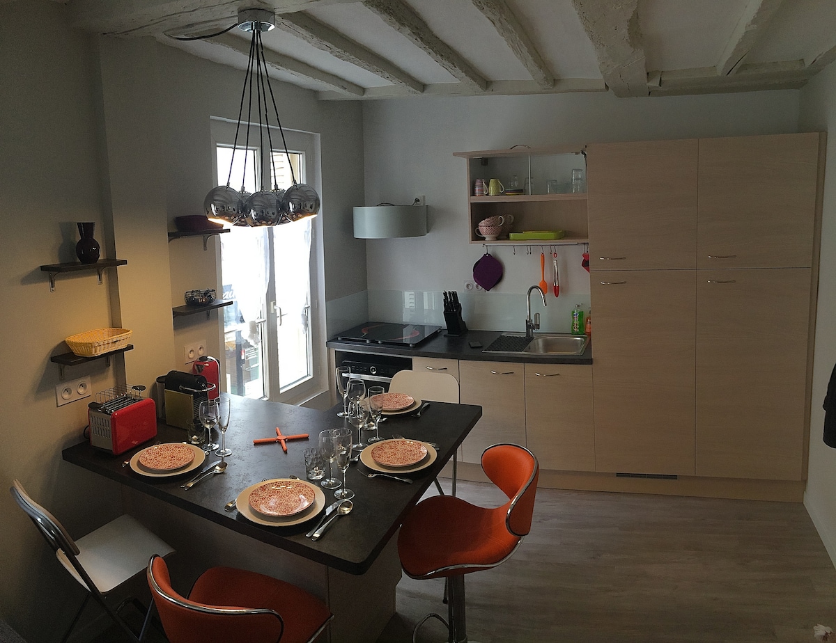 The compact kitchen features a dining table set for four, with elegant dishware and glassware arranged. Modern appliances include an induction cooktop, refrigerator, and microwave. Natural light enters through a nearby window, highlighting the clean lines of the cabinetry and the rustic ceiling beams.