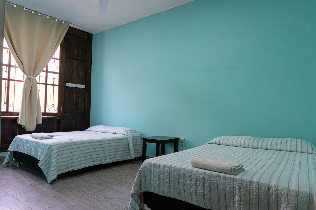 A room featuring two spacious beds with subtle striped bedding, positioned on a tiled floor. A dark wooden nightstand stands between them. Natural light enters through a large window, adorned with light curtains that partially frame the turquoise wall.