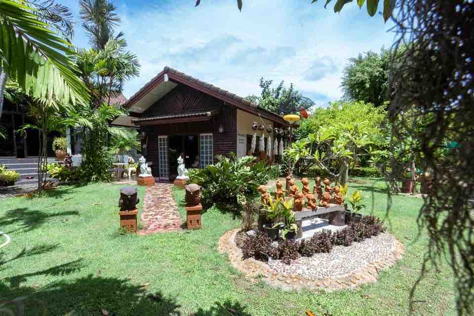A serene garden surrounds a wooden house, featuring a pathway lined with greenery and ornamental sculptures. The well-maintained lawn offers a variety of plants and trees, creating a tranquil atmosphere. In the foreground, a decorative stone arrangement complements the natural setting.