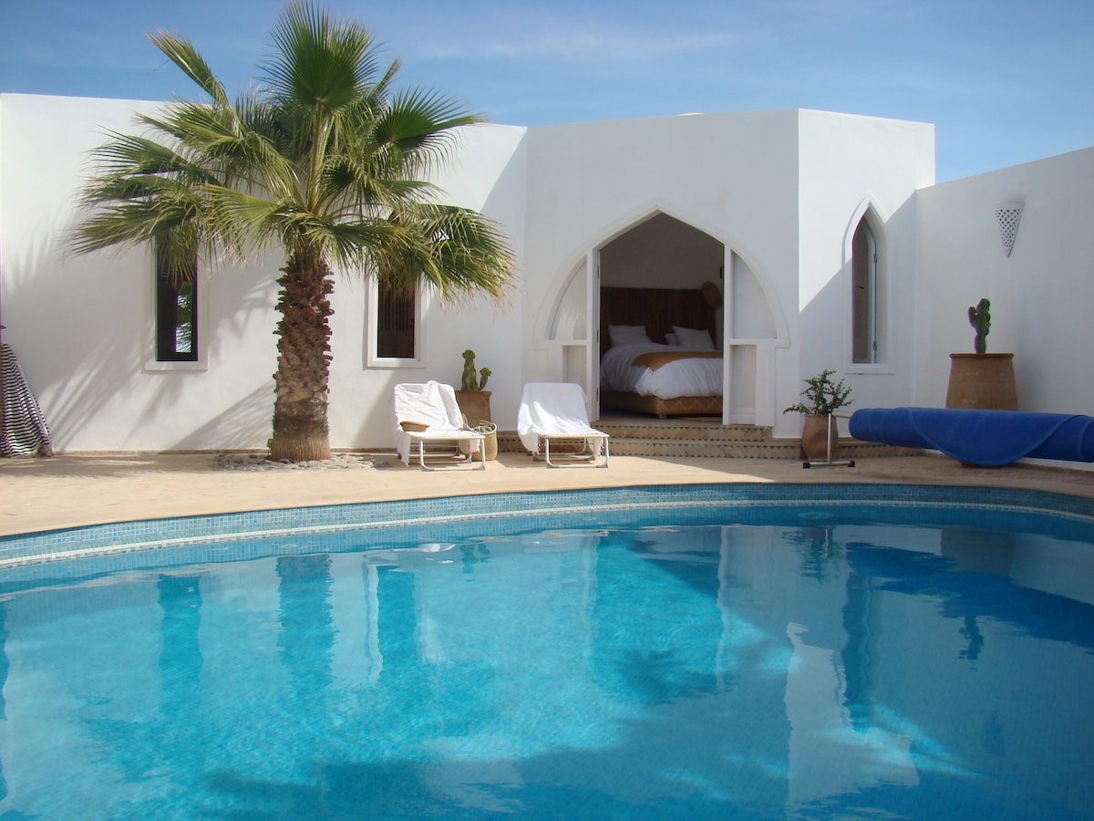 A serene outdoor area features a pool with clear blue water reflecting the sky. Two sun loungers are positioned beside the pool, while a palm tree provides shade. An arched opening reveals a spacious bed nestled in a cozy interior beyond.