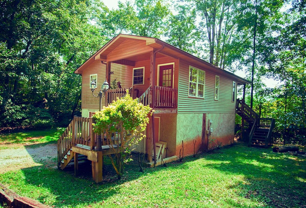 The exterior of the cabin is shown, featuring a light brown facade with large windows and a welcoming entryway. A staircase leads to the porch, surrounded by green lawns and trees. The space is designed for ease of access and blends into the natural surroundings.