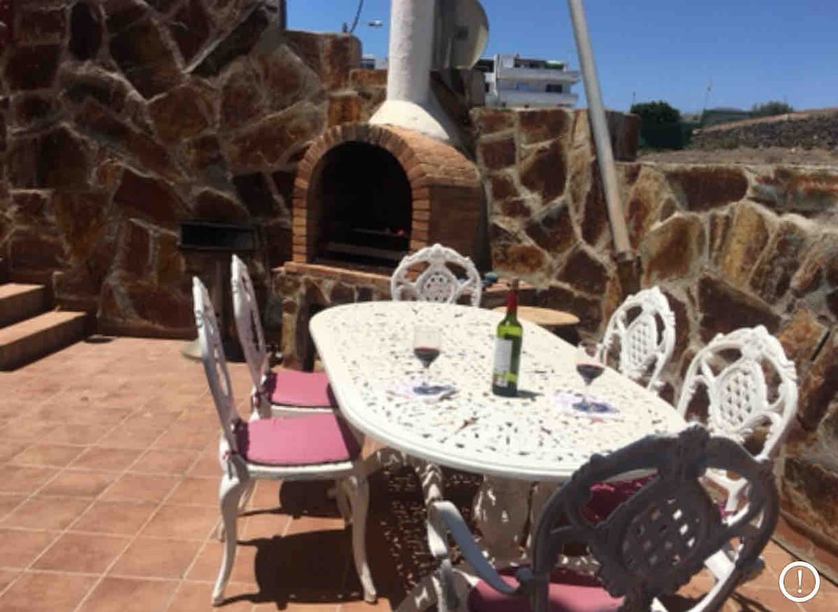 An outdoor dining area is set on a tiled terrace, featuring a large white table surrounded by six intricately designed chairs with soft cushions. A stone barbecue grill is visible in the background, along with a serene view of the surroundings and a bottle of wine at the table.