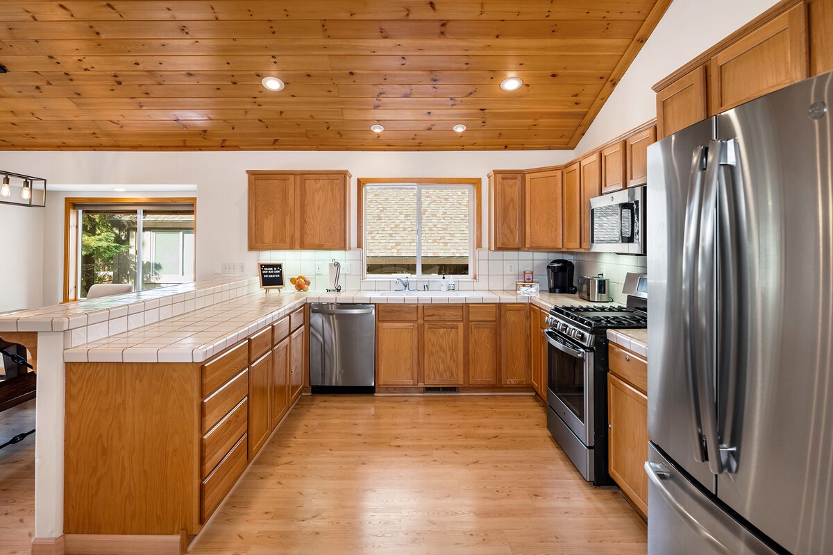 The spacious kitchen features warm wooden cabinetry and a tiled countertop, with stainless steel appliances including a gas stove and dishwasher. Natural light floods in from two windows, creating an inviting cooking space designed for comfort and functionality.