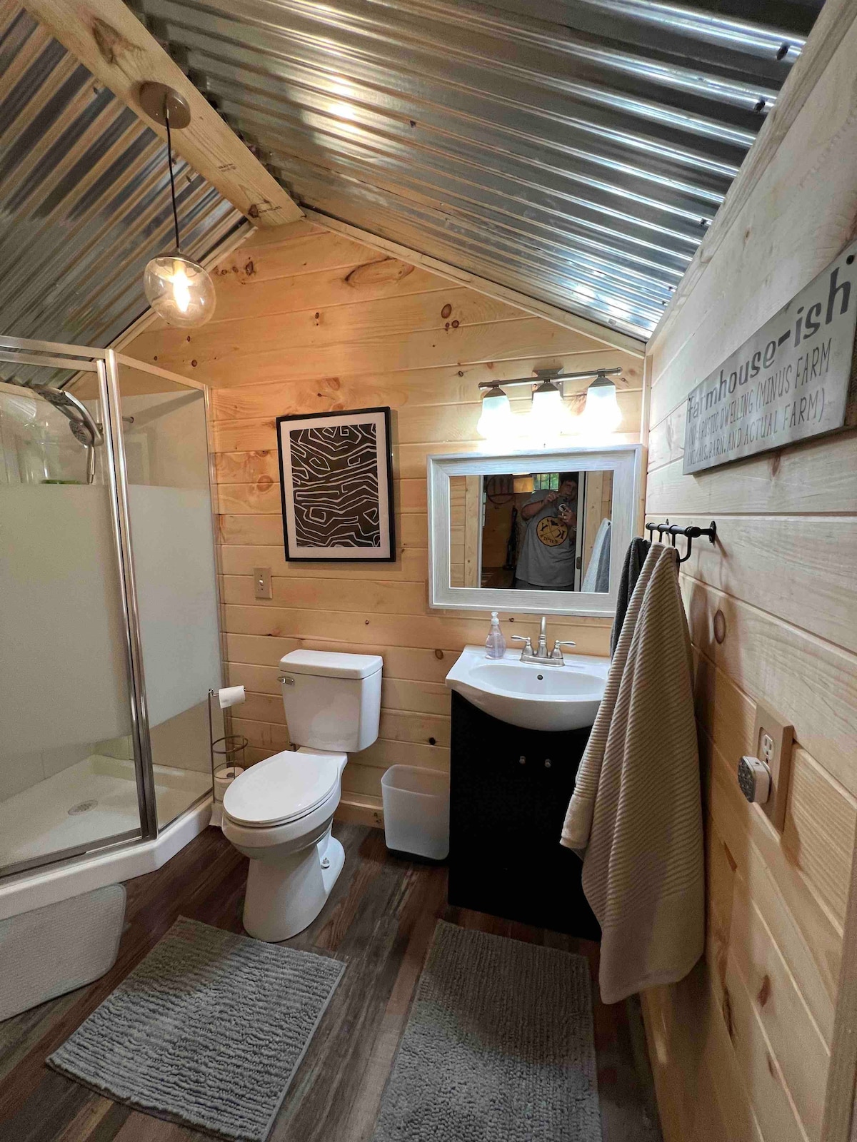 A compact bathroom features wooden walls and a corrugated metal ceiling. A glass shower enclosure stands to one side, with a modern sink and mirror opposite. A towel hangs on a bar, and two bath mats add comfort underfoot.