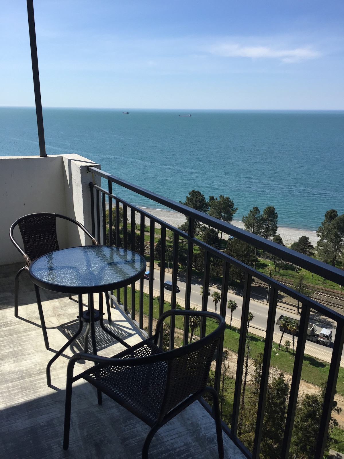 A balcony with two black woven chairs and a round glass table overlooks the sea. The expansive view includes ships gently floating on the water and a pathway lined with trees, enhancing the serene coastal atmosphere.