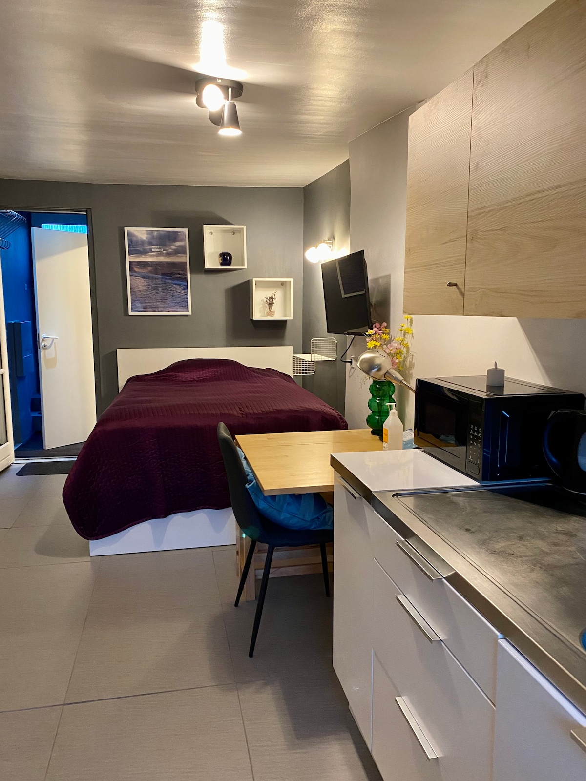A compact room includes a spacious bed dressed in a burgundy coverlet. A table with a green chair is positioned next to a kitchenette featuring a microwave and sink. A flat-screen TV is mounted on the wall, and a doorway leads to a bathroom area.