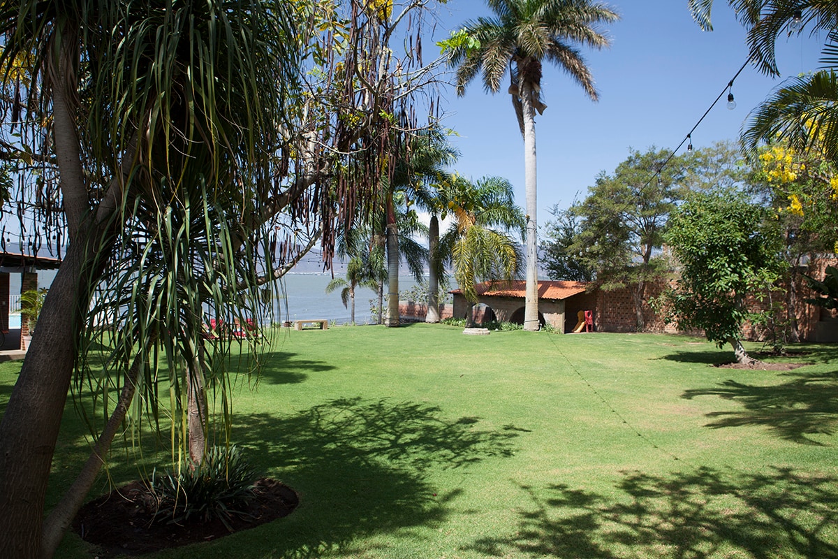 A spacious garden is displayed, featuring lush green grass and a variety of palm trees. The area extends towards the lake in the background, providing a serene natural setting. A shaded structure with a roof is visible in the distance.