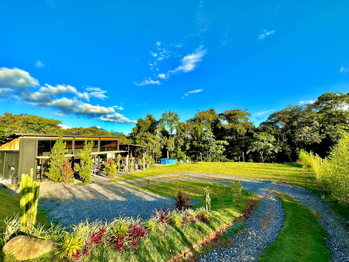 The exterior of the loft is surrounded by lush greenery, with a large grassy area visible. A gravel path leads towards the building, with colorful plants lining the edges. A swimming pool is situated to the side, and trees frame the idyllic scene under a clear blue sky.