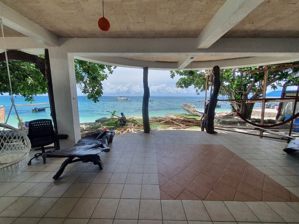 An open seating area features tiled flooring and shaded views of the beach. Comfortable lounge chairs are arranged for relaxation. The space is framed by lush greenery and provides direct sightlines to the inviting turquoise water, with boats gently floating in the distance.