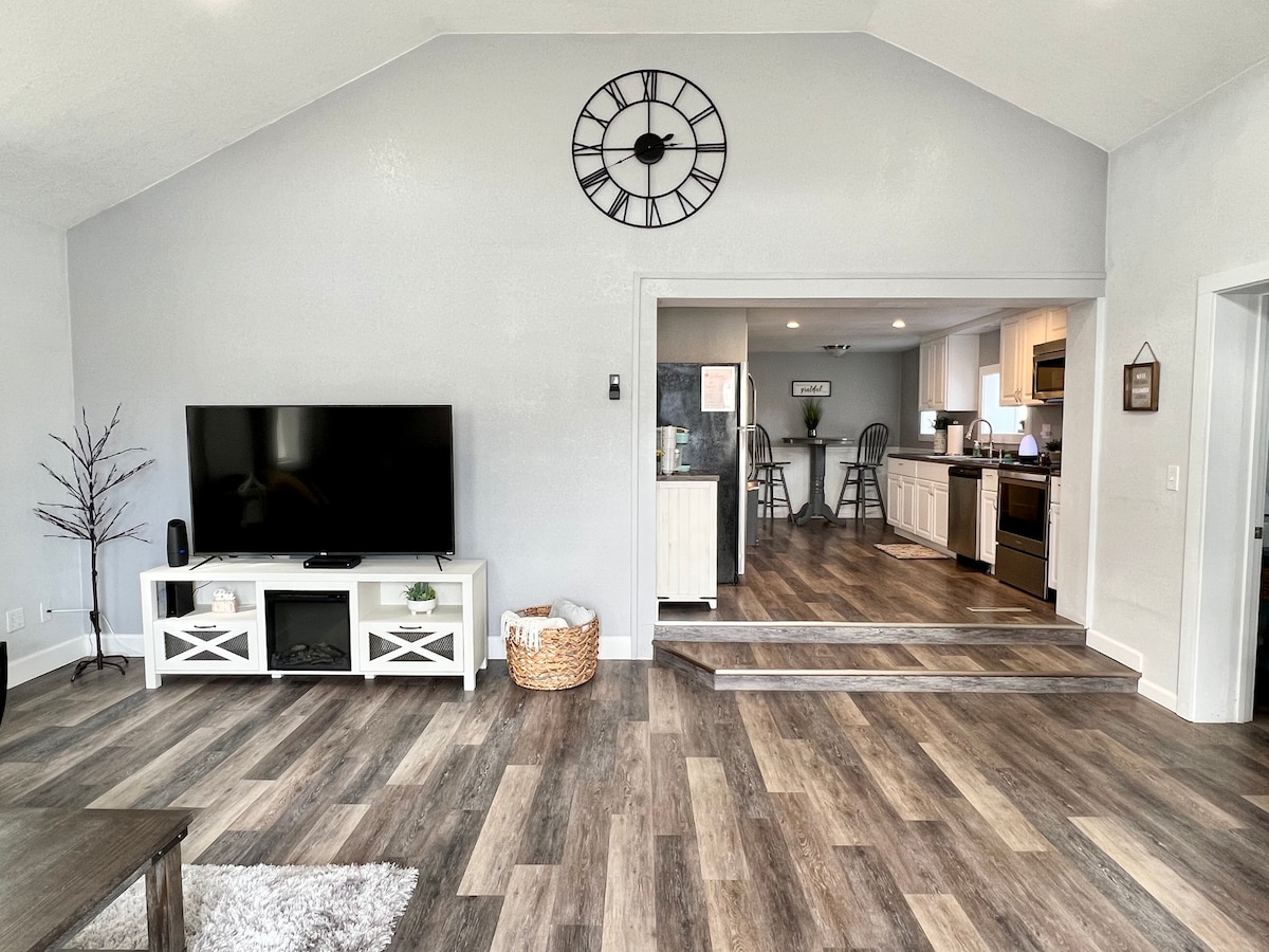 The living area features a modern design with a large flat-screen TV and a decorative clock above. Natural light floods the space, highlighting the open floor plan that leads to the kitchen. Two steps connect the living area to the kitchen for easy access.