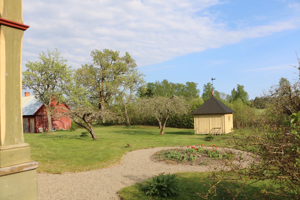 A spacious garden features a circular flower bed surrounded by green grass. Two apple trees are positioned nearby, with red barns visible in the background. A small yellow wooden gazebo adds a charming touch to the serene outdoor setting, under a partly cloudy sky.