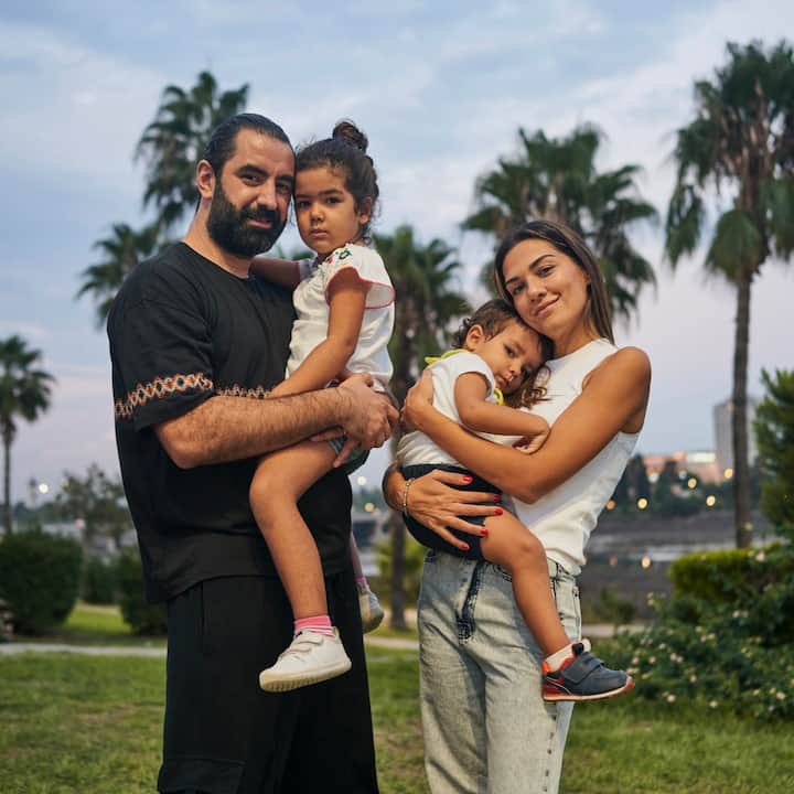 A father and mother smile serenely, holding their two children in a park with palm trees.