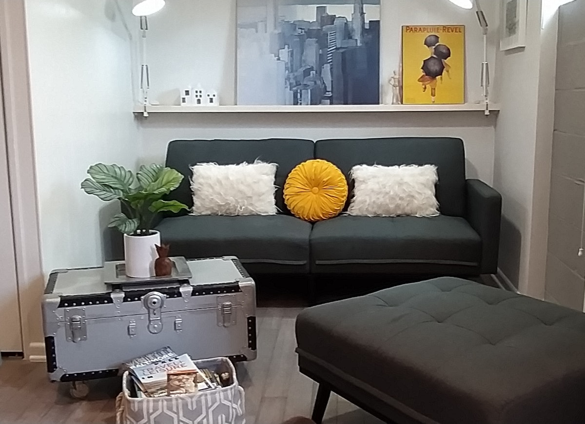A modern living area is presented with a dark green sofa adorned with fluffy pillows and a bright yellow accent cushion. A metal coffee table holds magazines, while a potted plant adds a touch of greenery. Artwork hangs on the wall above the sofa.