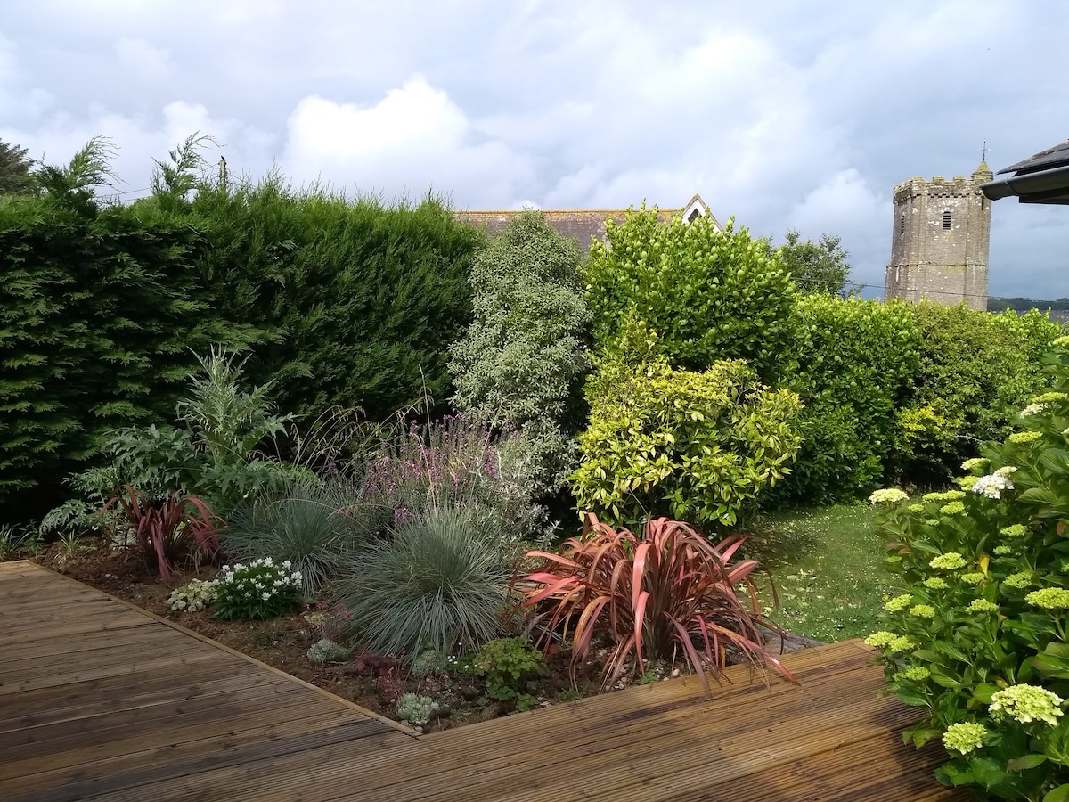 A lush garden area is presented with a wooden deck, showcasing a variety of plants and shrubs. The greenery includes ornamental grasses and flowering plants, with a tower from a nearby church visible in the background, adding character to the tranquil setting.