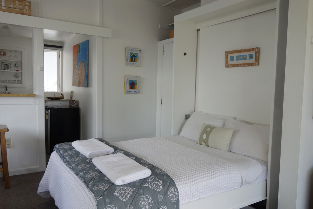 A spacious studio features a queen-size bed with a white quilt and decorative throws. Nearby, a kitchenette with minimalistic cabinetry is visible, alongside a fridge. Light is allowed in through a window draped with sheer curtains, enhancing the calm ambiance of the room.