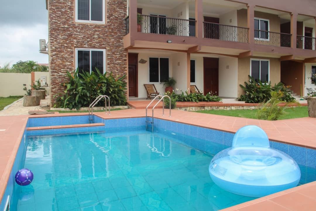 A swimming pool is surrounded by a charming patio. The clear blue water is complemented by a floating ring and a beach ball. Lush greenery borders the pool area, while the building features multiple balconies and large windows.