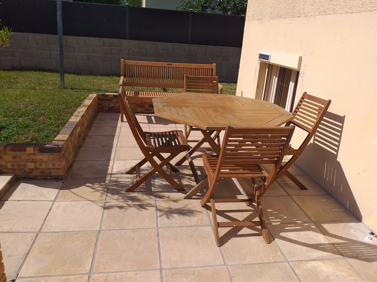 An outdoor patio area is depicted, featuring a round wooden table surrounded by five folding chairs. The patio has tiled flooring and is bordered by a brick wall. A patch of green grass is visible in the background, contributing to the serene outdoor setting.