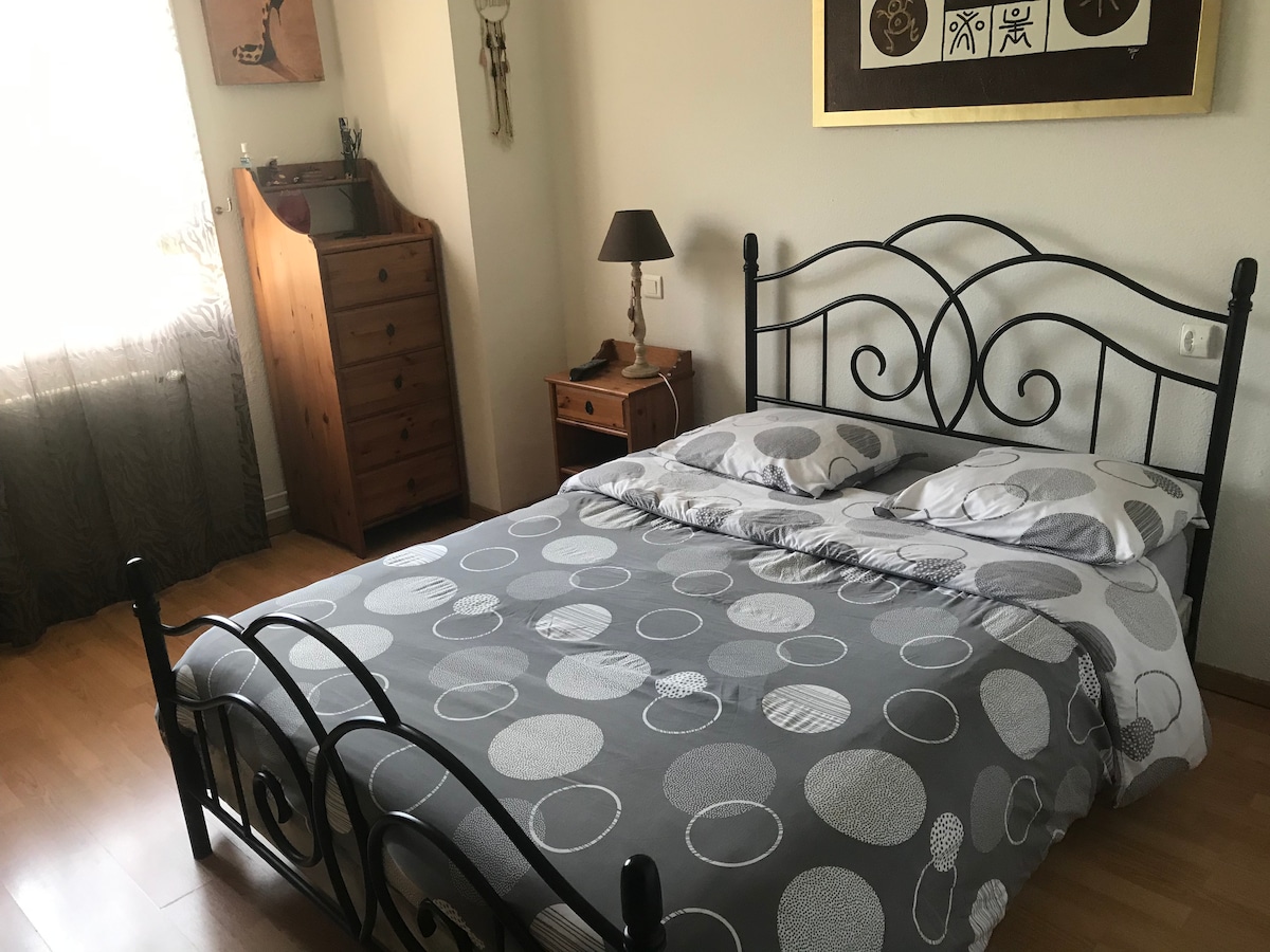 A bedroom with a graceful double bed featuring a patterned gray duvet and two pillows. A wooden dresser stands against one wall, complemented by a small bedside table with a lamp. Natural light filters through a nearby window, illuminating the inviting space.