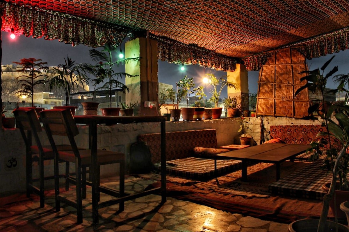 A serene outdoor seating area is adorned with potted plants, creating a natural ambiance. Traditional fabrics drape overhead, with soft lighting enhancing the space. Low tables and cushions invite relaxation, while natural textures and patterns contribute to the inviting design.
