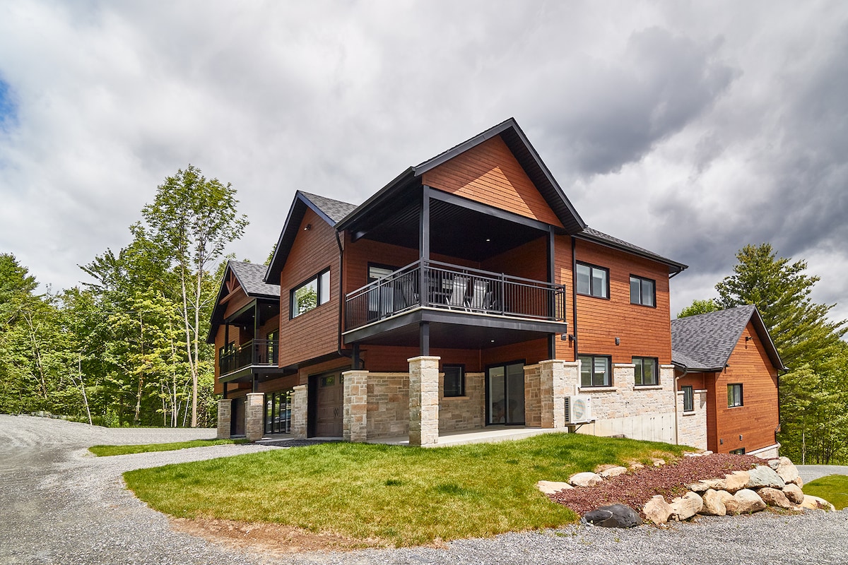 The two-story chalet is nestled in a natural setting, featuring a mix of wood and stone exterior. Large covered terraces are visible, offering outdoor seating. The driveway is gravel, surrounded by landscaped grass and strategically placed stones, enhancing the home's rustic appearance.