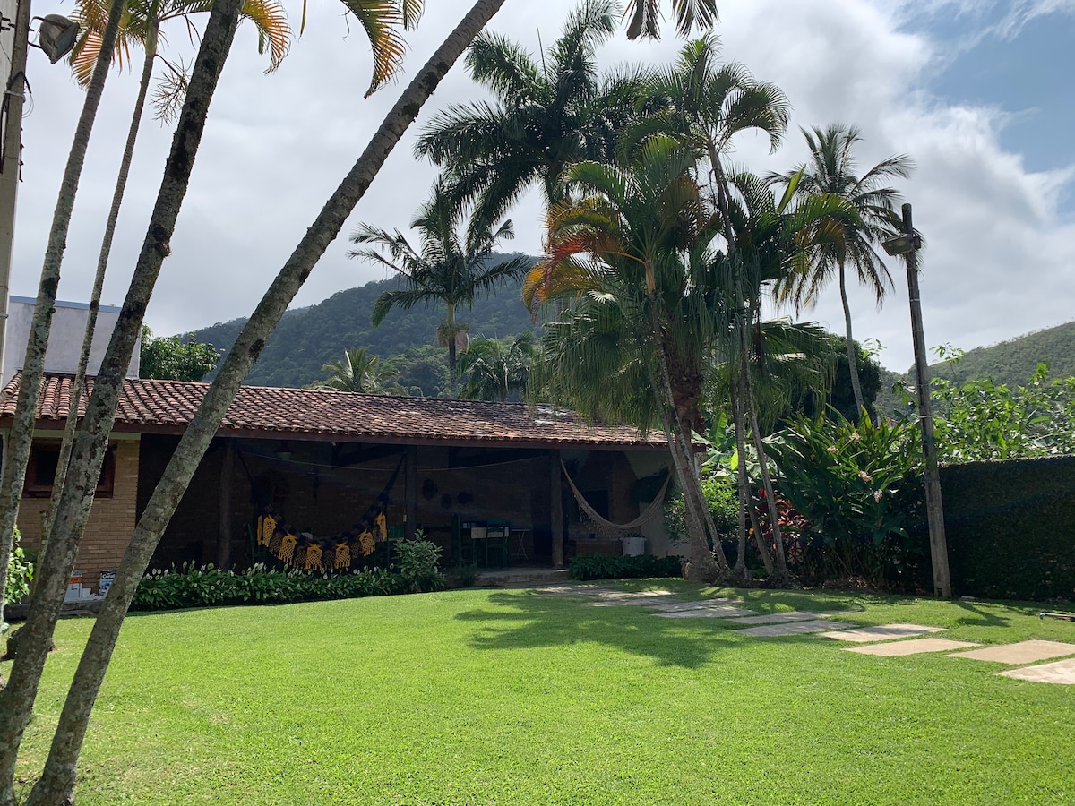 A spacious outdoor area is surrounded by lush greenery and palm trees. A well-maintained lawn features stone pathways leading to a covered patio. The patio is equipped with hammocks and a table, inviting relaxation in a tranquil setting with a mountain backdrop.