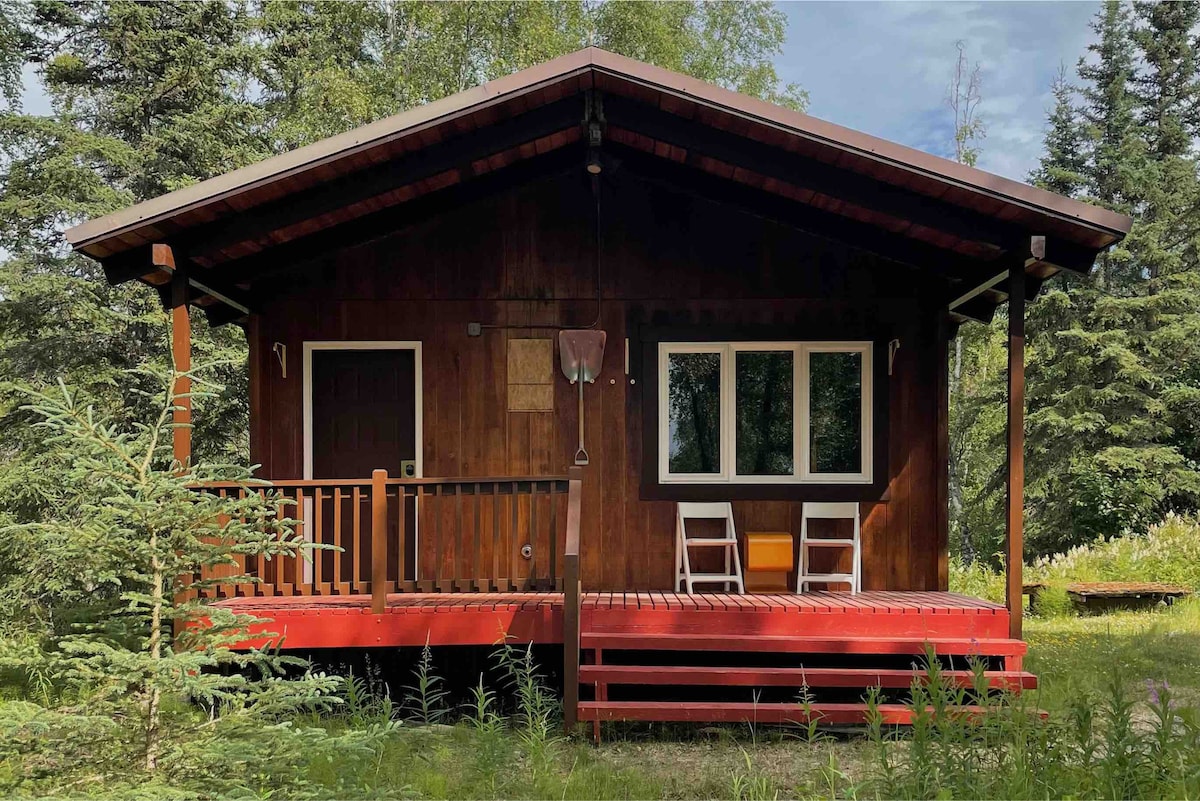 The exterior of a one-bedroom cabin features a rustic wooden facade and a welcoming porch. Two chairs are positioned on the deck, surrounded by lush greenery. Tall trees and a grassy area frame the cabin, creating a serene and natural setting.
