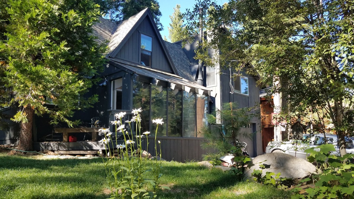 The exterior of a mountain home is set against a backdrop of tall trees. A sunroom with expansive windows extends from the side. A green lawn features wildflowers and boulders, creating a natural, inviting atmosphere around the property.