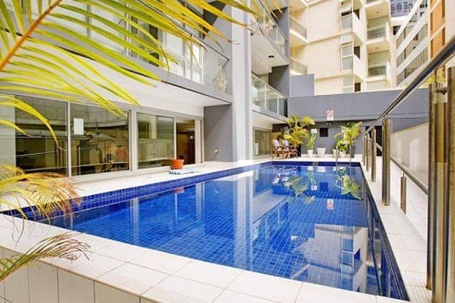 An outdoor heated swimming pool is framed by a tiled deck and surrounded by potted plants. Sunlight reflects off the water's surface, while lounge chairs are positioned nearby, offering a relaxing space for guests. The area is enclosed by modern condominium buildings.