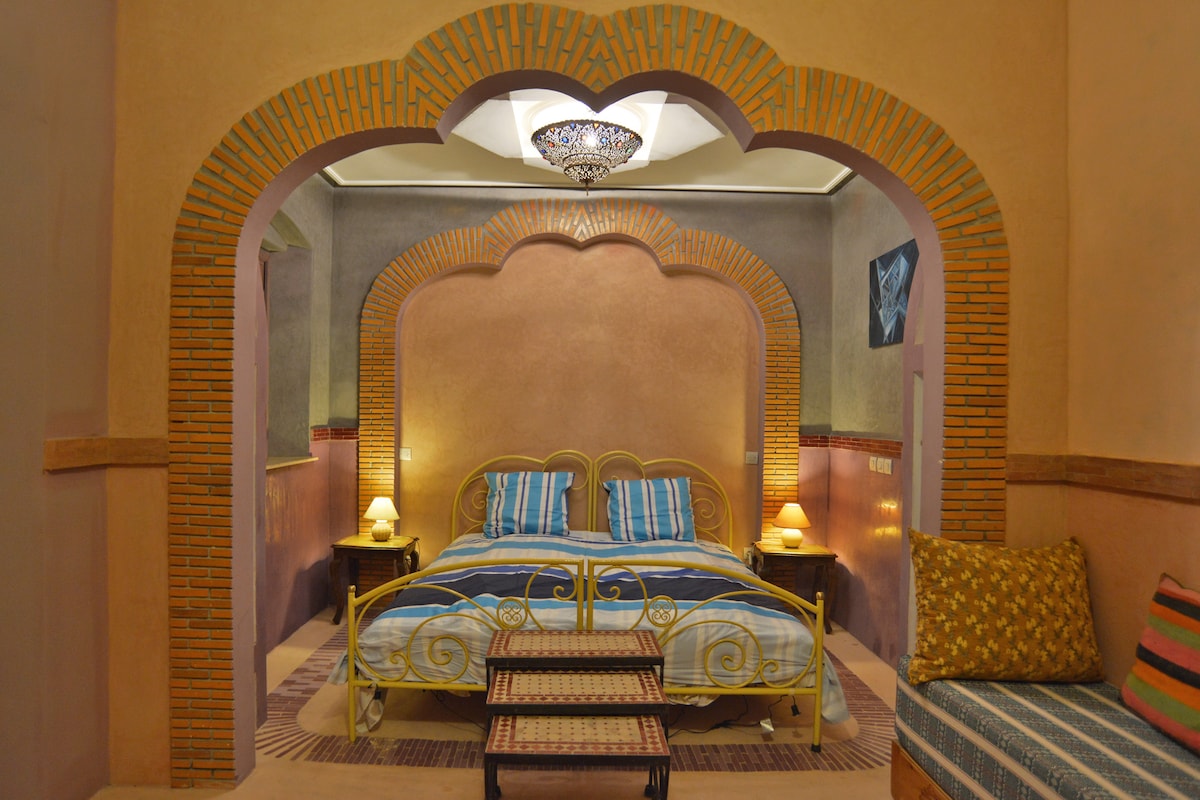 A spacious room is highlighted by an archway leading to a two-person bed with striped bedding. Nightstands with lamps are positioned on either side. Textured orange and brown walls complement the intricate ceiling light fixture. A seating area with colorful cushions is visible to the side.